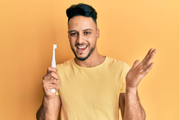 Junger arabischer Mann mit elektrischer Zahnbürste feiert Leistung mit glücklichem Lächeln und Siegerausdruck mit erhobener Hand  - Foto, Bild