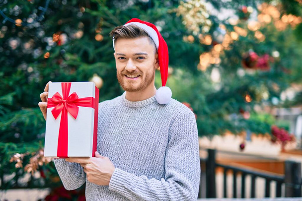 Joven hombre caucásico con sombrero de Navidad sosteniendo regalo en el parque. - Foto, imagen