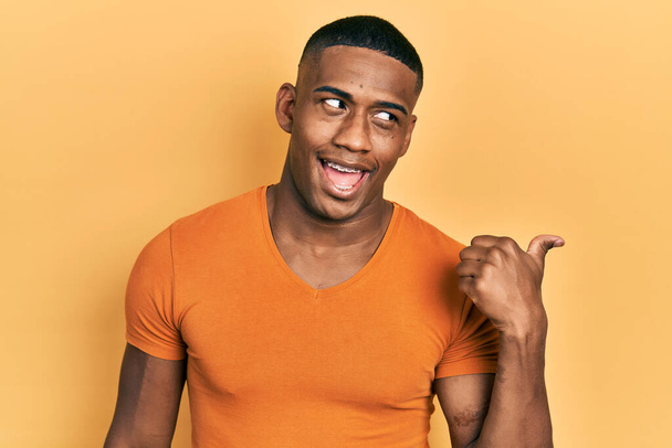 Fiatal fekete férfi alkalmi narancssárga pólót visel mosolygós boldog arccal néz és mutatva oldalra hüvelykujjával felfelé..  - Fotó, kép