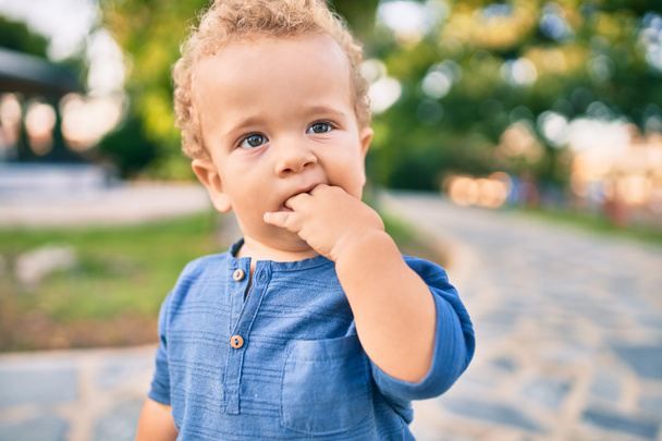 Sad little boy putting fingers on mouth touching gums because toothache at the park on a sunny day. Beautiful blonde hair male toddler in pain for new baby teeth outdoors - Zdjęcie, obraz