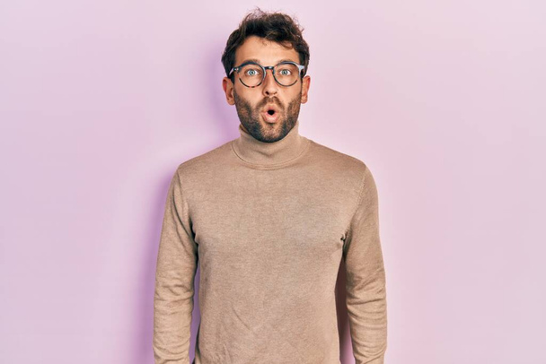 Handsome man with beard wearing turtleneck sweater and glasses afraid and shocked with surprise expression, fear and excited face.  - Photo, Image