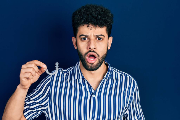 Jeune homme arabe avec barbe tenant invisible aligneur orthodontique et accolades peur et étonné avec la bouche ouverte pour surprise, visage incrédule  - Photo, image