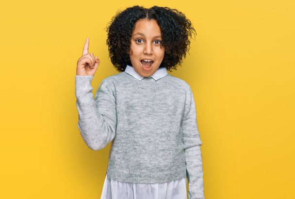 Kleines Mädchen mit Afro-Haaren in lässiger Kleidung, die mit erhobenem Zeigefinger auf eine erfolgreiche Idee hinweist. aufgeregt und glücklich. Nummer eins.  - Foto, Bild
