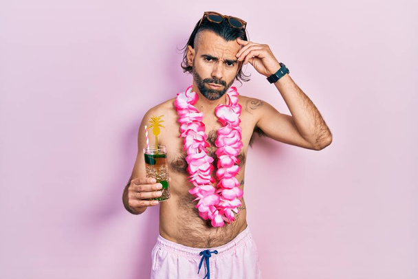 Joven hispano con traje de baño y lei hawaiano bebiendo cóctel tropical preocupado y estresado por un problema con la mano en la frente, nervioso y ansioso por la crisis  - Foto, imagen