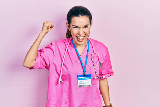 Young brunette woman wearing doctor uniform and stethoscope angry and mad raising fist frustrated and furious while shouting with anger. rage and aggressive concept.  - Photo, Image