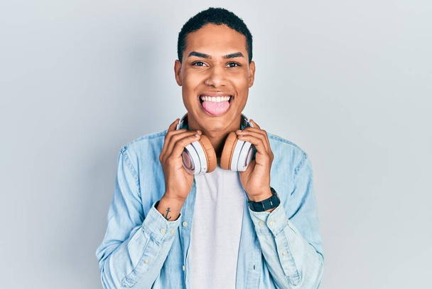 Młody Afroamerykanin słuchający muzyki za pomocą słuchawek wystaje język szczęśliwy z zabawnym wyrazem twarzy.  - Zdjęcie, obraz