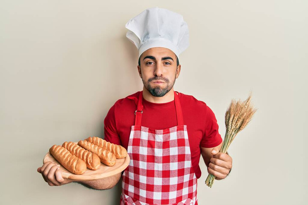 Jonge Spaanse man in bakkersuniform met zelfgemaakt brood en tarwepufwangen met een grappig gezicht. mond opgeblazen met lucht, vangen van lucht.  - Foto, afbeelding