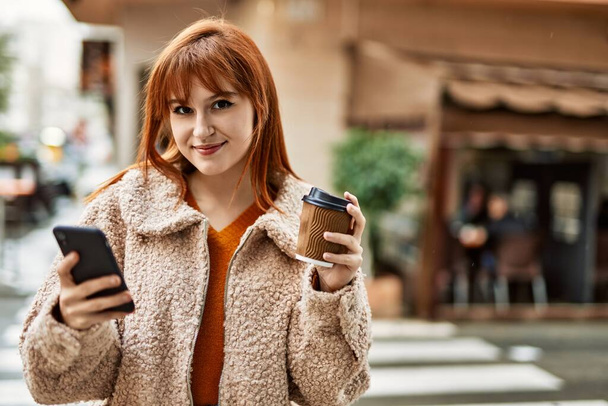 Young redhead girl smiling using smartphone drinking coffee at the city. - Foto, Imagem
