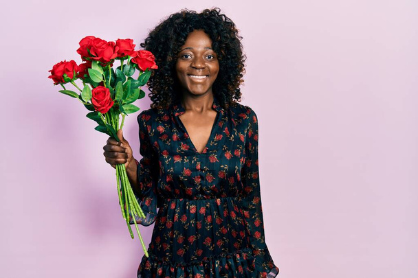 Junge afrikanisch-amerikanische Frau mit einem Strauß roter Rosen, die im Stehen positiv und glücklich aussehen und mit einem selbstbewussten Lächeln Zähne zeigen  - Foto, Bild