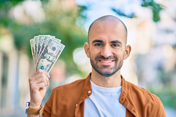 Mladý hispánec plešatý muž usměvavý šťastný drží nás dolary ve městě. - Fotografie, Obrázek