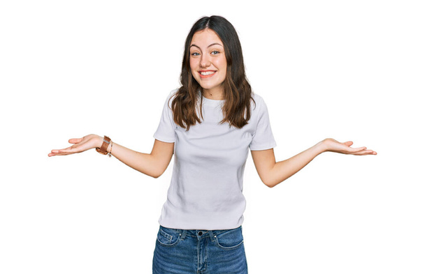 Jonge mooie vrouw draagt casual witte t-shirt glimlachen tonen beide handen open palmen, presenteren en reclame vergelijking en evenwicht  - Foto, afbeelding