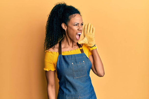 Middle age african american woman wearing professional apron shouting and screaming loud to side with hand on mouth. communication concept.  - Fotografie, Obrázek