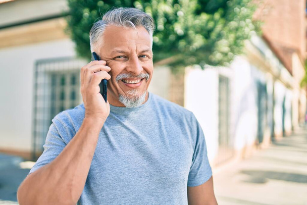 Hombre de pelo gris hispano de mediana edad sonriendo feliz hablando en el teléfono inteligente de la ciudad. - Foto, imagen