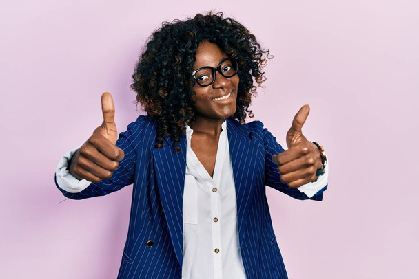 Jeune femme afro-américaine portant des vêtements de travail et des lunettes approuvant faire un geste positif avec la main, pouces levés souriant et heureux pour le succès. geste gagnant.  - Photo, image