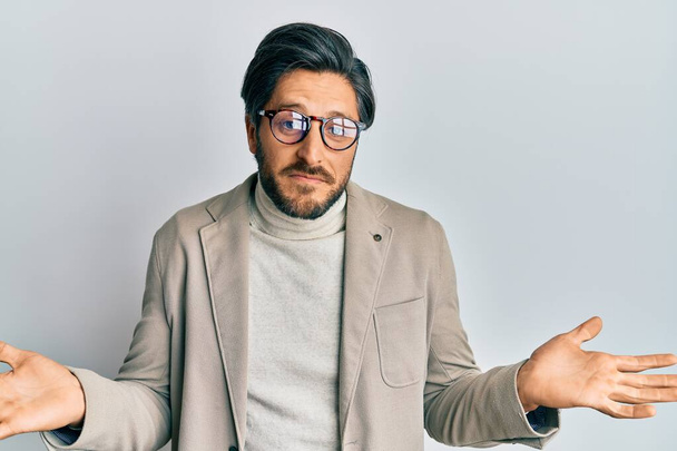 Young hispanic man wearing business jacket and glasses clueless and confused expression with arms and hands raised. doubt concept.  - Foto, Imagem