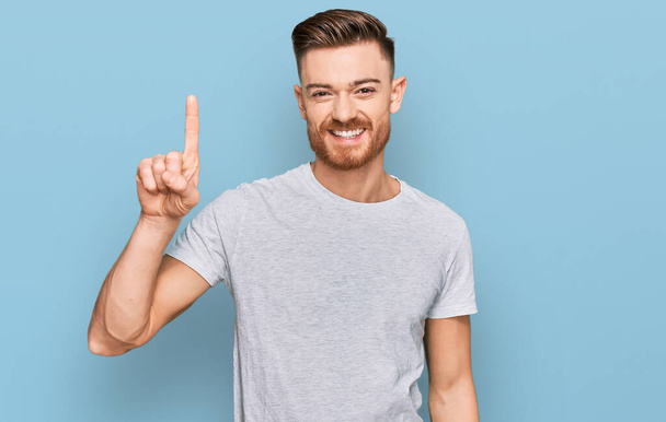 jovem ruiva homem vestindo casual cinza t-shirt mostrando e apontando para cima com o dedo número um enquanto sorrindo confiante e feliz.  - Foto, Imagem