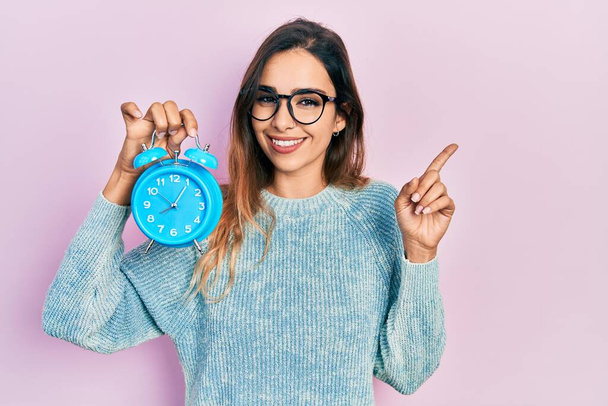 Young hispanic girl holding alarm clock smiling happy pointing with hand and finger to the side  - Photo, Image