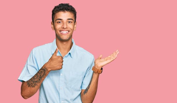 Jonge knappe Afro-Amerikaanse man draagt casual kleding met palmhand en doet ok gebaar met duimen omhoog, glimlachend gelukkig en vrolijk  - Foto, afbeelding