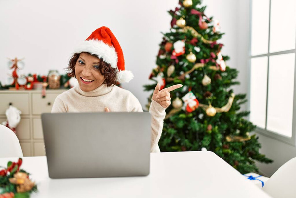 Middle age hispanic woman using laptop sitting by christmas tree smiling and looking at the camera pointing with two hands and fingers to the side.  - Photo, image