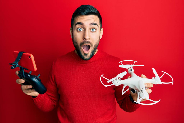 Νεαρός Ισπανόφωνος κρατώντας drone και τηλεχειριστήριο με smartphone φοβισμένος και σοκαρισμένος από έκπληξη και κατάπληκτος έκφραση, φόβο και ενθουσιασμένο πρόσωπο.  - Φωτογραφία, εικόνα