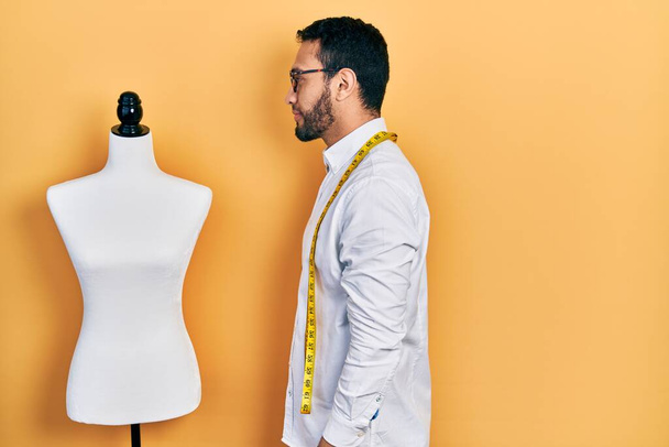 Hispanic man with beard dressmaker designer standing by manikin looking to side, relax profile pose with natural face with confident smile.  - Foto, Bild