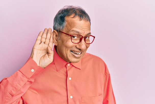 Keski-ikäinen intialainen mies päällään rento vaatteita ja lasit hymyillen käsi korvan yli kuunnellen huhua tai juoruja. kuurous käsite.  - Valokuva, kuva