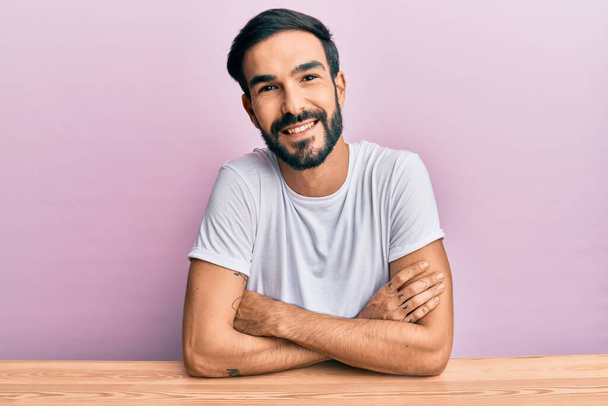 Junger hispanischer Mann in lässiger Kleidung sitzt auf dem Tisch und lächelt glücklich mit verschränkten Armen in die Kamera. positive person.  - Foto, Bild