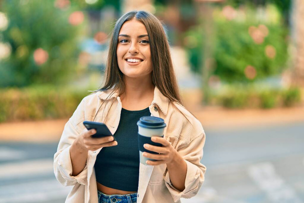 Młoda latynoska dziewczyna uśmiecha się szczęśliwy za pomocą smartfona i picia zabrać kawę w parku. - Zdjęcie, obraz