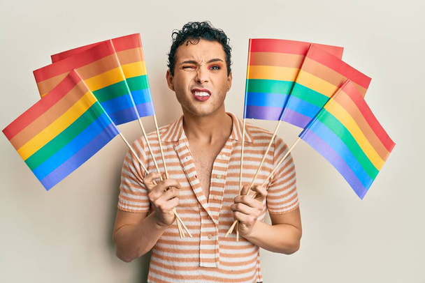Hombre guapo usando maquillaje sosteniendo banderas de arco iris lgbtq expresión despistada y confusa. concepto de duda.  - Foto, imagen
