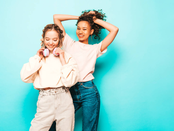 Two young beautiful smiling international hipster female in trendy summer clothes. Sexy carefree women posing near blue wall in studio. Positive models having fun. Concept of friendship - 写真・画像
