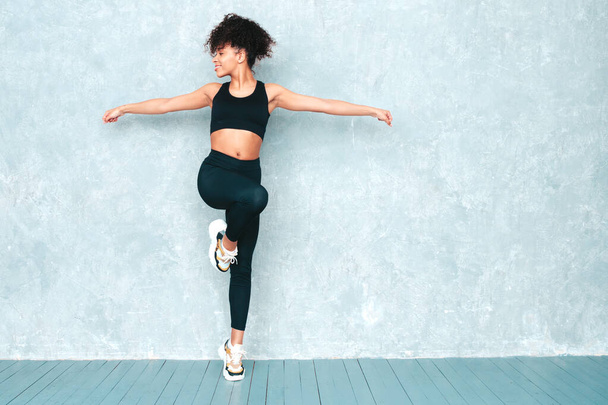 Fitness smiling black woman in sports clothing with afro curls hairstyle.She wearing sportswear. Young beautiful model with perfect tanned body.Female jumping and running in studio near gray wall - Foto, immagini