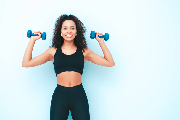 Fitness smiling black woman in sports clothing with afro curls hairstyle.She wearing sportswear. Young beautiful model with perfect tanned body.Female holding dumbbells in studio near light blue wall - Foto, Bild