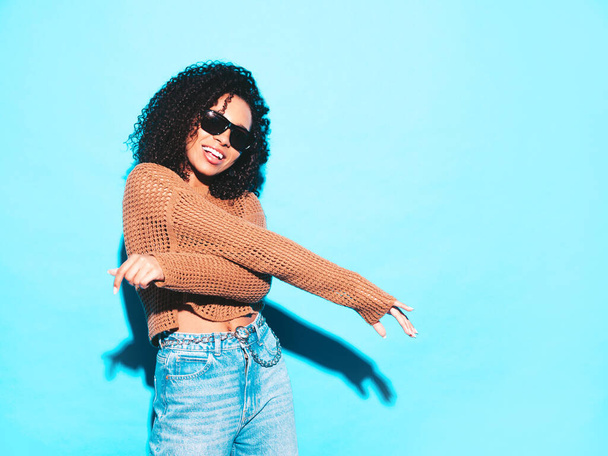 Beautiful black woman with afro curls hairstyle.Smiling model in trendy summer clothes. Sexy carefree female posing near blue wall in studio. Tanned and cheerful in sunglasses. Dancing hip hop  - Foto, immagini