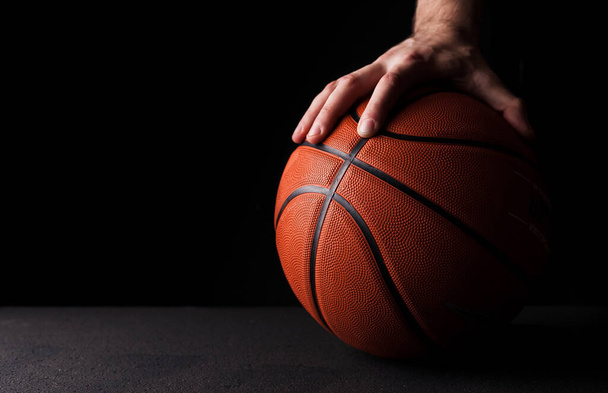 Rubber orange basketball ball is holding by a hand on a black background. - Фото, изображение