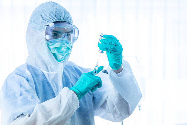 Koncept vakcinace COVID-19. Asijský lékař s lékařskou rukavicí, maskou a skafandrem PPE drží injekční stříkačku a vakcínu proti viru Corona. - Fotografie, Obrázek