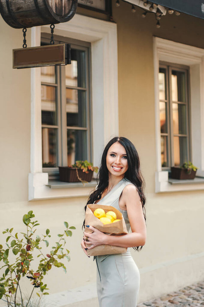 Eine schöne, gebräunte, sexy südliche Schönheit steht auf einem kunterbunten Hintergrund, umgeben von Palmblättern und hält eine hölzerne Schale mit Zitronen in ihren Händen. - Foto, Bild