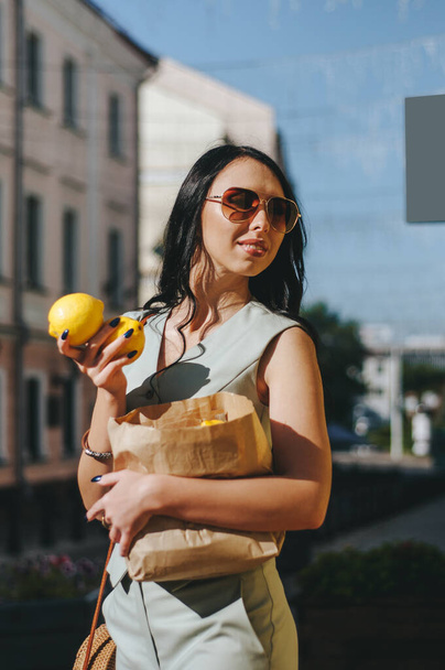 Young beautiful stylish woman wearing green dress, black jeans jacket and sunglasses walking at the street in city. Pretty brunette girl with long hair holding paper bag with lemons - Fotoğraf, Görsel