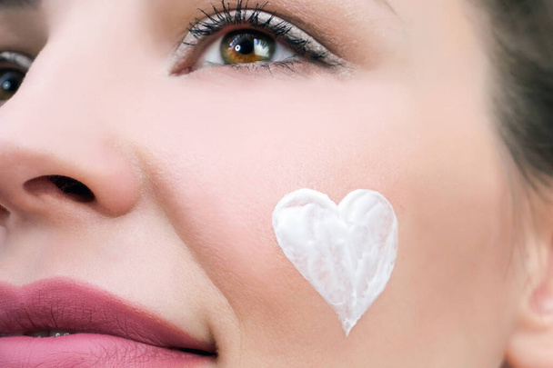 Facial skin care concept. A young woman has a heart of cream on her face. - Foto, Imagen