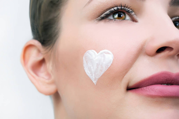 Facial skin care concept. A young woman has a heart of cream on her face. - Foto, Bild