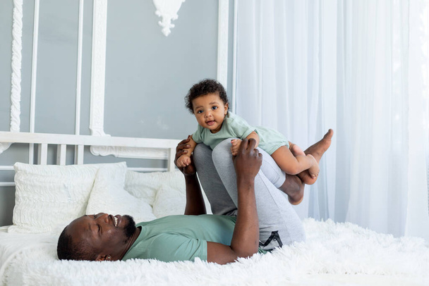 счастливый улыбающийся африканский американский папа с маленьким сыном на кровати дома обниматься и играть, счастливая семья, День отца - Фото, изображение