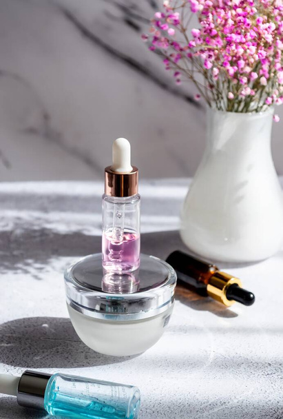 Skin care and beauty concept. Cosmetics different serums and cream on a light marble background, shadows, flowers in a vase, place for text. - Photo, Image