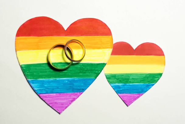 Dos anillos dorados en los corazones recortados de papel coloreados como una bandera LGBT, color blanco en el fondo, enfoque selectivo. concepto de corazón para lesbianas, gays, bisexuales, transgénero y personas queer matrimonio y unión - Foto, Imagen