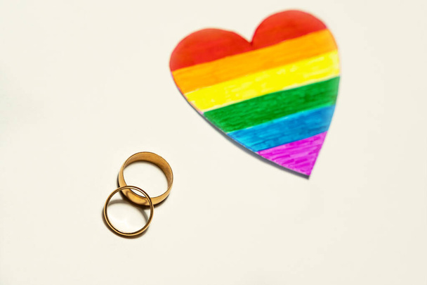 Zwei goldene Ringe und das Herz aus Papierausschnitt in der Farbe der LGBT-Flagge. Herzkonzept für lesbische, schwule, bisexuelle, transsexuelle und queere Menschen - Foto, Bild