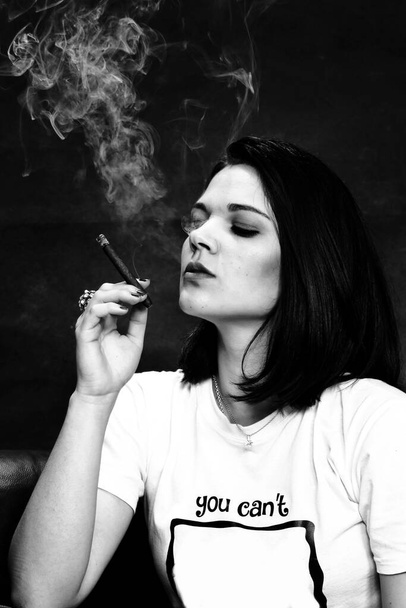 Όμορφη ευρωπαϊκή αυταρχική μελαχρινή κοπέλα καπνίζει cigarillo σαν αφεντικό σε στούντιο σε απομονωμένο φόντο. Μοντελοποίηση, στυλ, έννοια της μόδας. Επίδραση θορύβου - Φωτογραφία, εικόνα