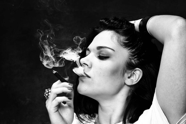 Schöne europäische bossy brünettes Mädchen raucht einen Zigarillo wie ein Chef im Studio auf isoliertem Hintergrund. Modeln, Stil, Modekonzept. Noir-Effekt - Foto, Bild