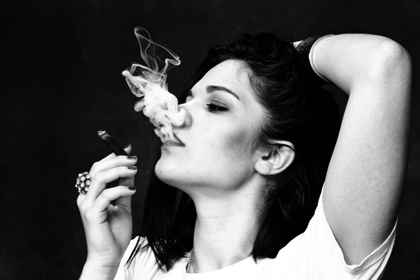 Όμορφη ευρωπαϊκή αυταρχική μελαχρινή κοπέλα καπνίζει cigarillo σαν αφεντικό σε στούντιο σε απομονωμένο φόντο. Μοντελοποίηση, στυλ, έννοια της μόδας. Επίδραση θορύβου - Φωτογραφία, εικόνα