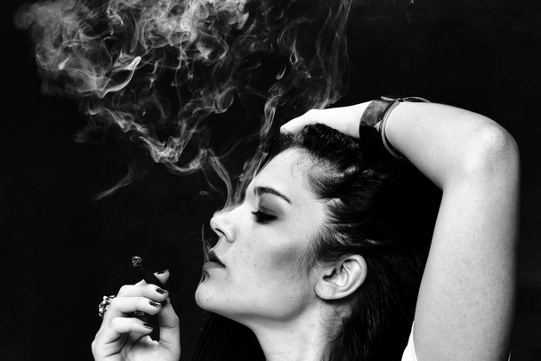 Piękna europejska apodyktyczna brunetka paląca cygaretki jak szef w studio na odosobnionym tle. Modelowanie, styl, koncepcja mody. Efekt noir - Zdjęcie, obraz