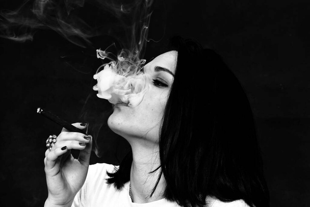 Hermosa chica morena mandona europea fumando un cigarrillo como un jefe en el estudio sobre un fondo aislado. Modelado, estilo, concepto de moda. Efecto negro - Foto, imagen