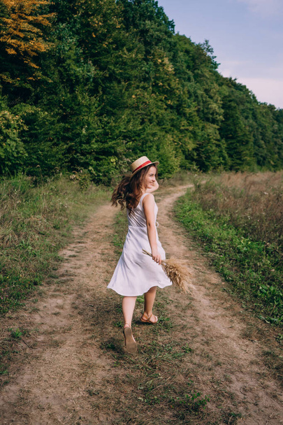 Молодая женщина в платье с букетом бежит по полю. Улыбающаяся девушка в шляпе бежит по тропе теплым летним днем. - Фото, изображение