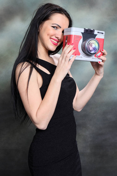 Menina europeia bonita com cabelo preto longo posando em estúdio sobre tela de fundo e segurando uma câmera falsa. Estilo, tendências, conceito de moda. - Foto, Imagem